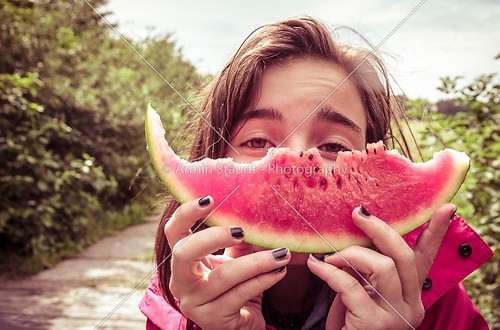 girl is holding a bitten slice of melon in front of here face, v