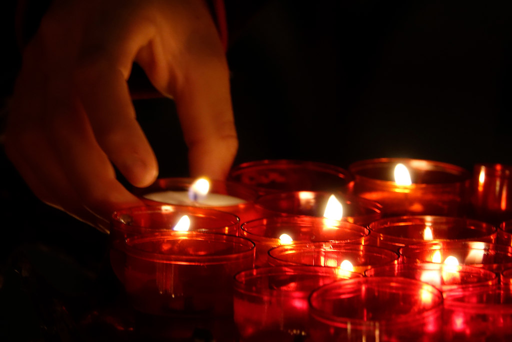 Memory (Memory) | human hand lights candles in a church | glowing, flame, candle, burning, illuminated, tea light, one person, real people, indoors, human hand, lit, close-up, night, human body part, church